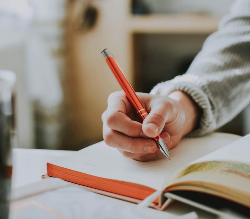 pexels-photo-1925536-1925536 A person writing in a notebook with a red ballpoint pen, showcasing detail and focus.